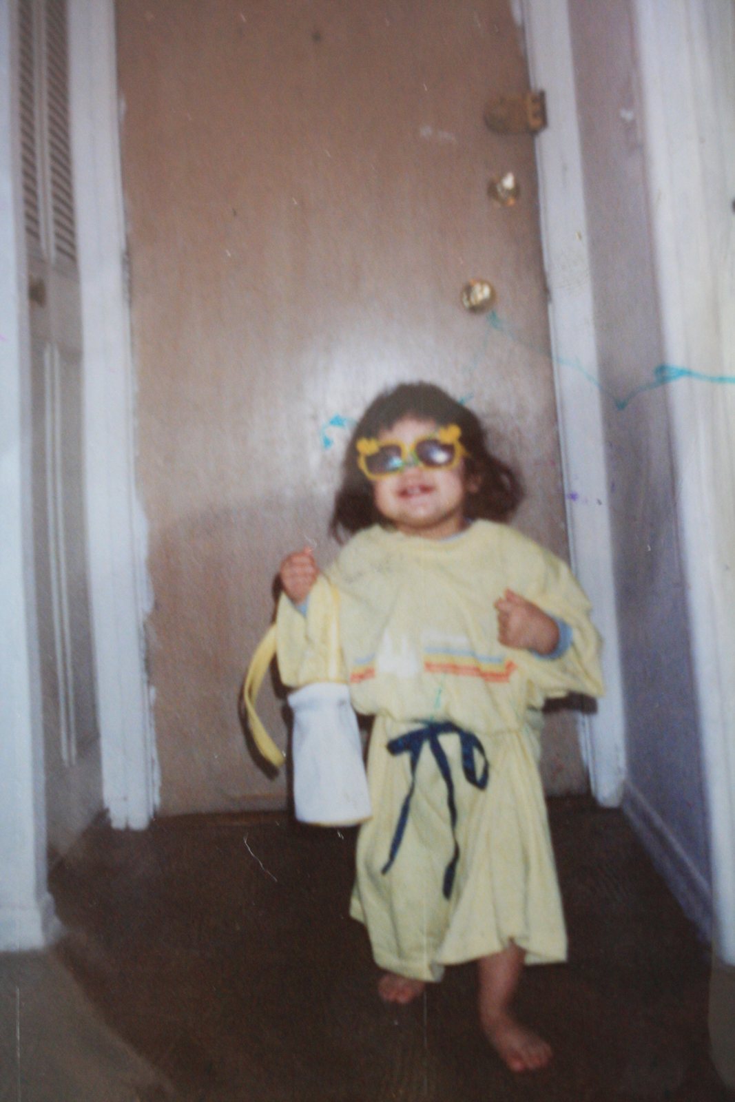 Shereen as a toddler in oversized sunglasses and a yellow dress, hallway at home